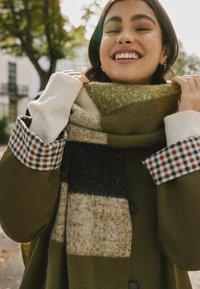 Écharpe en laine avec des rayures vertes, noires et crèmes, tenue par une personne portant un manteau olive avec un poignet intérieur à carreaux et des manches blanches.