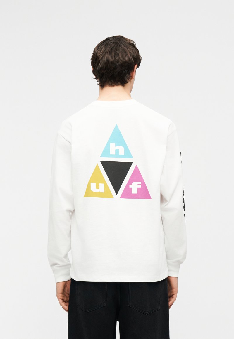 Άτομο με κοντά σκούρα μαλλιά φοράει λευκό πουκάμισο με μακριά μανίκια που δείχνει τρία πολύχρωμα τρίγωνα (μπλε, κίτρινο, ροζ) με τα γράμματα "h", "u" και "f" στην πλάτη.