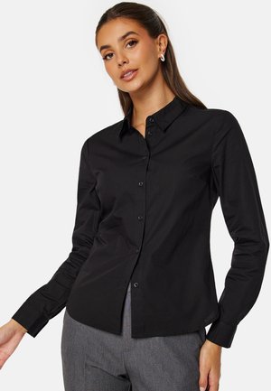Chemise noire à manches longues en tissu lisse, avec un design à boutons sur le devant, un col pointu et une coupe ajustée.