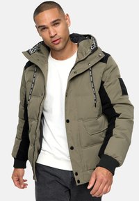 Doudoune vert olive avec accents noirs, fermeture éclair à l'avant, boutons-pression, poignets côtelés et deux poches latérales. La capuche est dotée de cordons de serrage de marque.