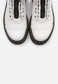 Zapatos de cuero blancos con suela negra y costuras amarillas, que cuentan con un cierre de cremallera y un diseño de cordones tradicional en la parte superior.