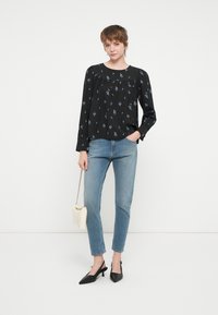 Svart langermet bluse med blå blomstermønster, sammen med lyseblå jeans. Modellen holder en hvit veske og har på seg svarte spisse hæler.