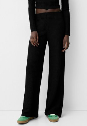 Personne portant un pantalon noir texturé à jambes larges, un crop top noir à manches longues, et des baskets vertes avec semelles en gomme, debout devant un fond blanc.