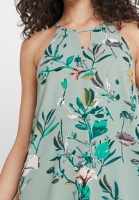 Ärmlös blus i mintgrön med nyckelhålsknäppning och blommönster i nyanser av grönt, rosa och grått. Mjuk och lätt tyger.