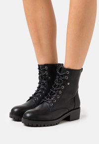 Botas de cuero negro hasta el tobillo con cordones y ojales de metal, acabado texturizado, tacón cuadrado, cremallera lateral, suela de goma duradera y sin patrones visibles.