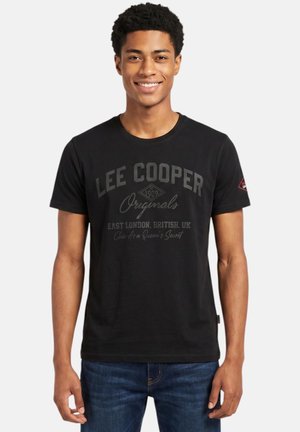 Jeune homme souriant, portant un t-shirt noir Lee Cooper Originals avec texte et un jean bleu, debout sur un fond blanc uni.