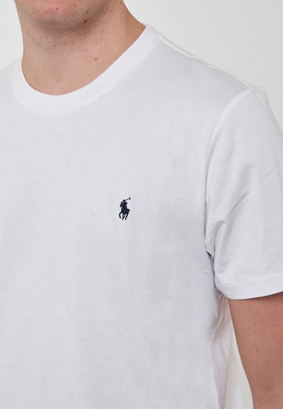 Maglietta a maniche corte in cotone bianco con scollo rotondo, presenta un piccolo logo blu navy di un cavallo e un cavaliere nella parte sinistra del torace. Texture morbida e design minimale.