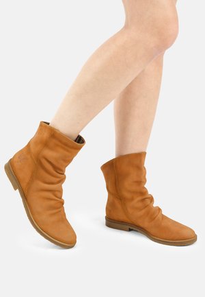 Piernas con botas de cuero marrón suave hasta la mitad de la pantorrilla con suelas planas, un pie ligeramente levantado sobre un fondo claro y liso.