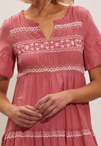 Vaaleanpunainen puuvillamekko, jossa on lyhyet hihat, V-kaula-aukko ja hienovaraisia valkoisia brodeerauksia rinnassa ja helmassa. Laskostettu muotoilu, jossa on tekstuuria.
