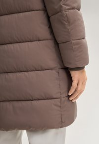 Giacca puffa marrone con texture trapuntata, caratterizzata da maniche aderenti e polsini elastici. La mano appoggiata lungo la cucitura aggiunge dettaglio al design.