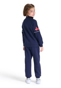 Tuta blu navy con colletto alto e accenti bianchi a contrasto. Presenta un logo rosso sulla manica sinistra e pantaloni affusolati.