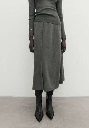 Personne portant un haut gris à manches longues, une jupe midi grise assortie, des bottes à talons noires à bout pointu, debout devant un fond uni.