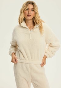 Kremowy polarowy pulower z wysokim kołnierzem, pół zamkiem błyskawicznym i elastycznym pasem; charakteryzuje się miękką, puszystą teksturą i oversizowymi rękawami.