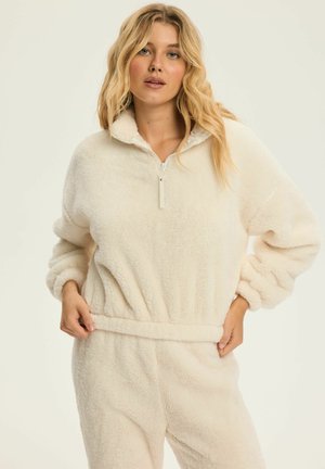 Kremowy polarowy pulower z wysokim kołnierzem, pół zamkiem błyskawicznym i elastycznym pasem; charakteryzuje się miękką, puszystą teksturą i oversizowymi rękawami.