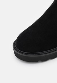 Schwarze Wildleder-Ankle-Boots mit runder Zehenform, strukturiertem Finish und einer dicken Gummisohle, die am Rand mit einer genähten Akzentuierung versehen ist.