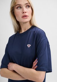 Mörkblå bomullst-shirt med korta ärmar och rund halsringning; har en liten, broderad logotyp på bröstet. Slät textur.