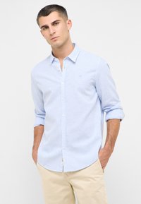 Camicetta button-up a righe azzurro chiaro con maniche lunghe, caratterizzata da un logo discreto e una trama morbida, abbinata a pantaloni corti color crema.