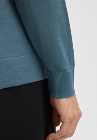 Hellblaues Sweatshirt mit gerippten Bündchen und Saum, weicher Textur und leichtem Glanz, taillierte Passform, die den Arm und die Hand zeigt.