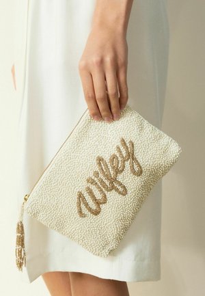 Mână care ține o geantă plic albă cu mărgele și cuvântul "wifey" din mărgele aurii, fermoar cu ciucure, pe fundal de rochie albă.