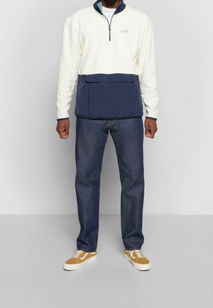 Pull-over crème et marine avec une demi-fermeture éclair, doté d'une grande poche frontale. Associé à un jean bleu foncé et des baskets jaunes.