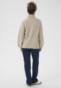 Beige gestrickter Pullover mit hohem Kragen, der ein Zopfmuster aufweist. Trägt dazu dunkelblaue Hosen und hellfarbige Sneakers.