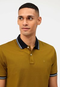 Olivegrünes Poloshirt mit einem marineblauen Kragen mit weißen Streifen, dreiknöpfiger Knopfleiste und einem kleinen Logo auf der linken Brust. Strukturierter Stoff.