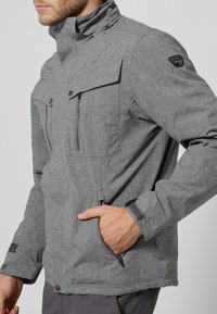 Graue Outdoor-Jacke mit hohem Kragen, zwei Brusttaschen, durchgehendem Reißverschluss und strukturiertem Material. Mit Logodetail auf der Schulter.