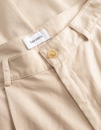 Οι μπεζ βαμβακερές παντελόνες διαθέτουν φαρδύ λάστιχο στη μέση με κλείσιμο κουμπιού και ετικέτα που αναγράφει "LES DEUX". Το ύφασμα έχει υφή με ριγωτό σχέδιο.