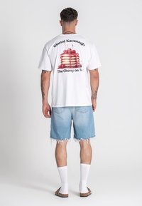 Camiseta blanca con un gráfico de un pastel en capas decorado con cerezas rojas y el texto "Gianni Kavanagh The Cherry on Top", combinada con shorts de mezclilla.