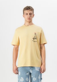 Ung mand iført en lysegul t-shirt med et flamingo-bird grafik og tekst på brystet, parret med blå mønstrede shorts.