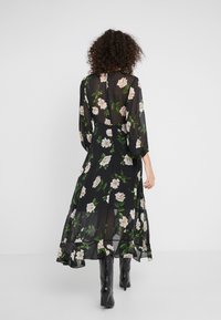 Zwarte bloemen maxi-jurk met doorschijnende stof, lange pofmouwen en een vloeiende rok. Het heeft witte bloemen met groene bladeren en een sleutelgatrug.