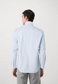 Camisa de botones a rayas azul claro con cuello clásico, mangas largas y puños con botones amarillos. Se lleva con pantalones grises.
