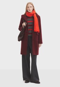 Cappotto di lana borgogna con cappuccio, abbinato a un maglione a righe grigie e rosse. Gli accessori includono una sciarpa arancione e una borsa marrone scuro, indossati con pantaloni a righe.