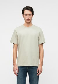 Light gray t-shirt in soft fabric, short sleeves, crew neck, and a minimal logo on the chest. Paired with dark blue jeans.