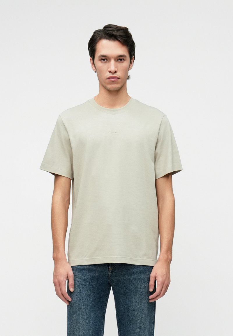 Light gray t-shirt in soft fabric, short sleeves, crew neck, and a minimal logo on the chest. Paired with dark blue jeans.