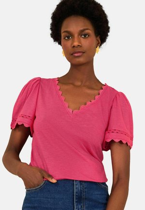 Femeie cu păr scurt și creț, purtând un top roz cu decolteu în V valurit, mâneci bufante și blugi bleumarin, cu mâna sprijinită în buzunar.