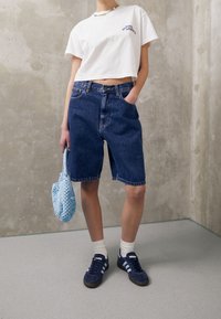 Camiseta blanca recortada con logo, pantalones cortos de mezclilla azul, bolso azul tejido al crochet, calcetines blancos y zapatillas azul marino con rayas blancas.