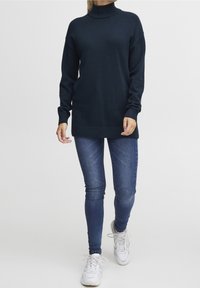 Ciemnogranatowy sweter z golfem, długimi rękawami, luźnym krojem i prążkowanymi mankietami, zestawiony z dopasowanymi niebieskimi dżinsami i białymi trampkami.