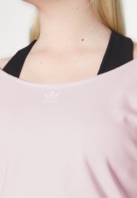 Camisa rosa clara com um decote largo e mangas curtas, apresentando uma alça atlética preta por baixo e um pequeno logo branco da Adidas no peito.