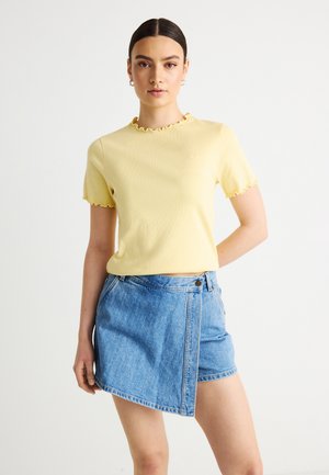 Žena oblečená v žltej blúzke s krátkym rukávom s volánikmi a asymetrickej modrej rifľovej mini sukni na bielom pozadí.