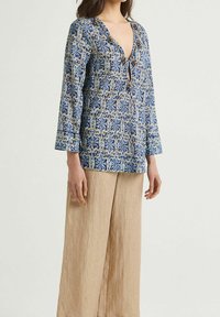 Tunic blu e crema con un motivo, scollatura a V e dettagli decorativi in perline, abbinata a pantaloni larghi in lino beige.