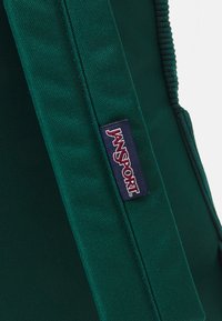 Green Jansport backpack detail featuring textured fabric, padded straps, and a visible logo tag stitched onto the side.