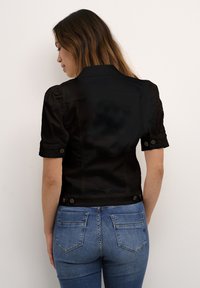 Svart kort jacka med korta puffärmar, knappdetaljer och en slät textur, bärs över åtsittande blå jeans.