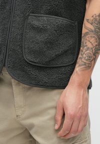 Pilkas fliso liemenė su tekstūruota paviršiumi, turi didelę kišenę su juodu apvadu ir kontrastingą užtrauktuką.