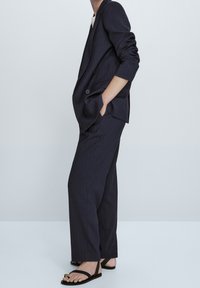 Personne portant un costume bleu marine à fines rayures avec les manches retroussées et des sandales noires, debout les mains dans les poches devant un fond uni.