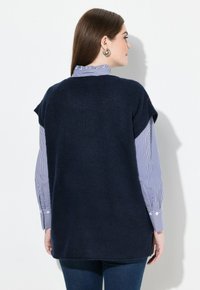 Žena na sobě má námořnickou modrou bezrukávovou svetr přes modro-bíle pruhovanou košili s knoflíky, stojí bokem proti bílé zdi.