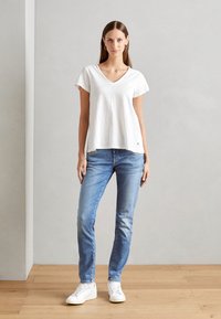 Weißes kurzärmliges V-Neck T-Shirt mit lockerem Schnitt, kombiniert mit hellblauen Jeans und weißen Sneakers. Einfaches Design ohne sichtbare Muster.