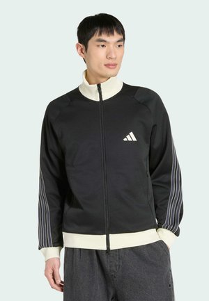 Giovane uomo che indossa una giacca nera con zip, colletto e polsini bianchi, maniche a righe bianche e logo Adidas sul petto.