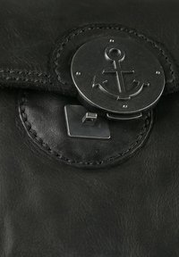 Bolso de cuero negro con un broche redondo de ancla plateada, con costuras detalladas y un acabado texturizado. El broche es metálico y geométrico.