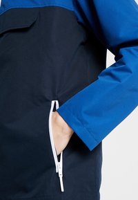 Navy and blue jacket featuring a zippered side pocket, smooth texture, and contrast color blocking at the shoulders. White zipper detailing.